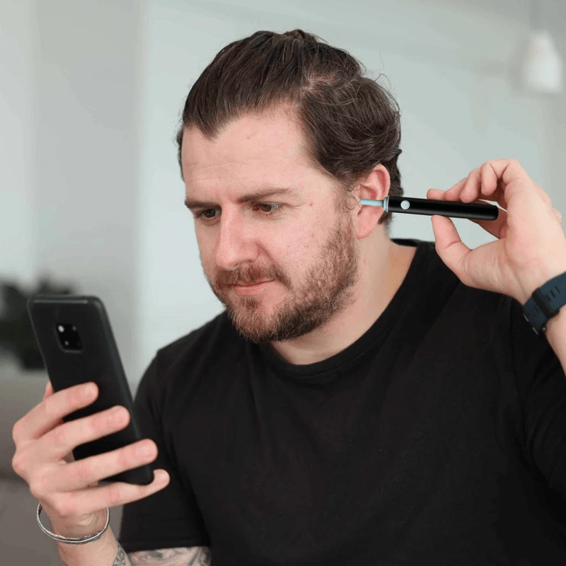 Man using a Smart Ear Cleaner to safely clean his ears while looking at a smartphone – effective and safe ear cleaning device